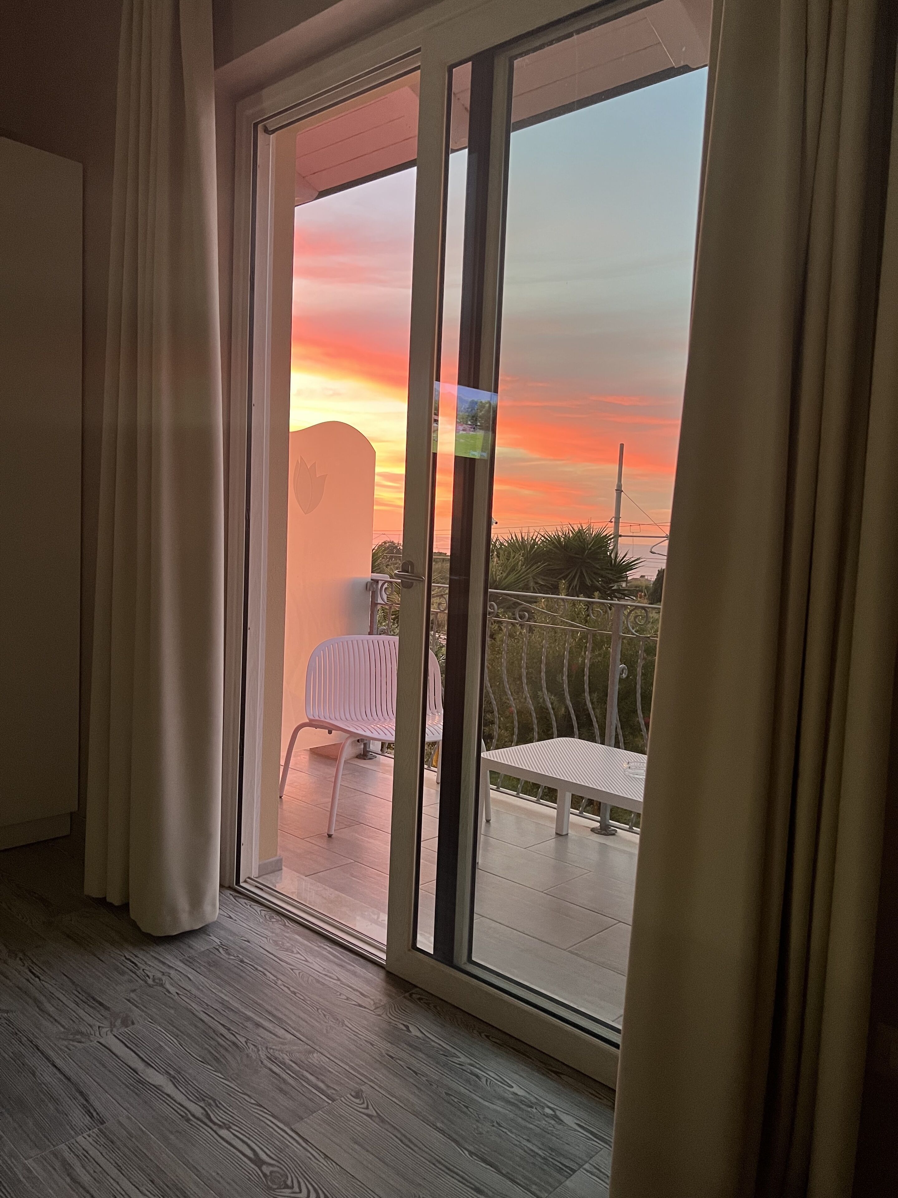 Una habitación con puerta de vidrio corredera que da a un balcón exterior con vistas a un atardecer.