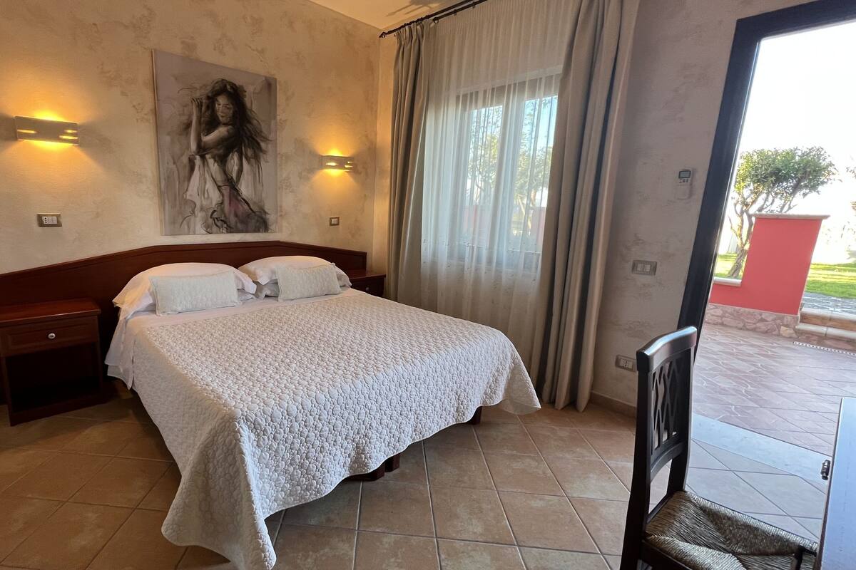 A hotel room with a bed, nightstands, a painting, and a view of the outdoors.