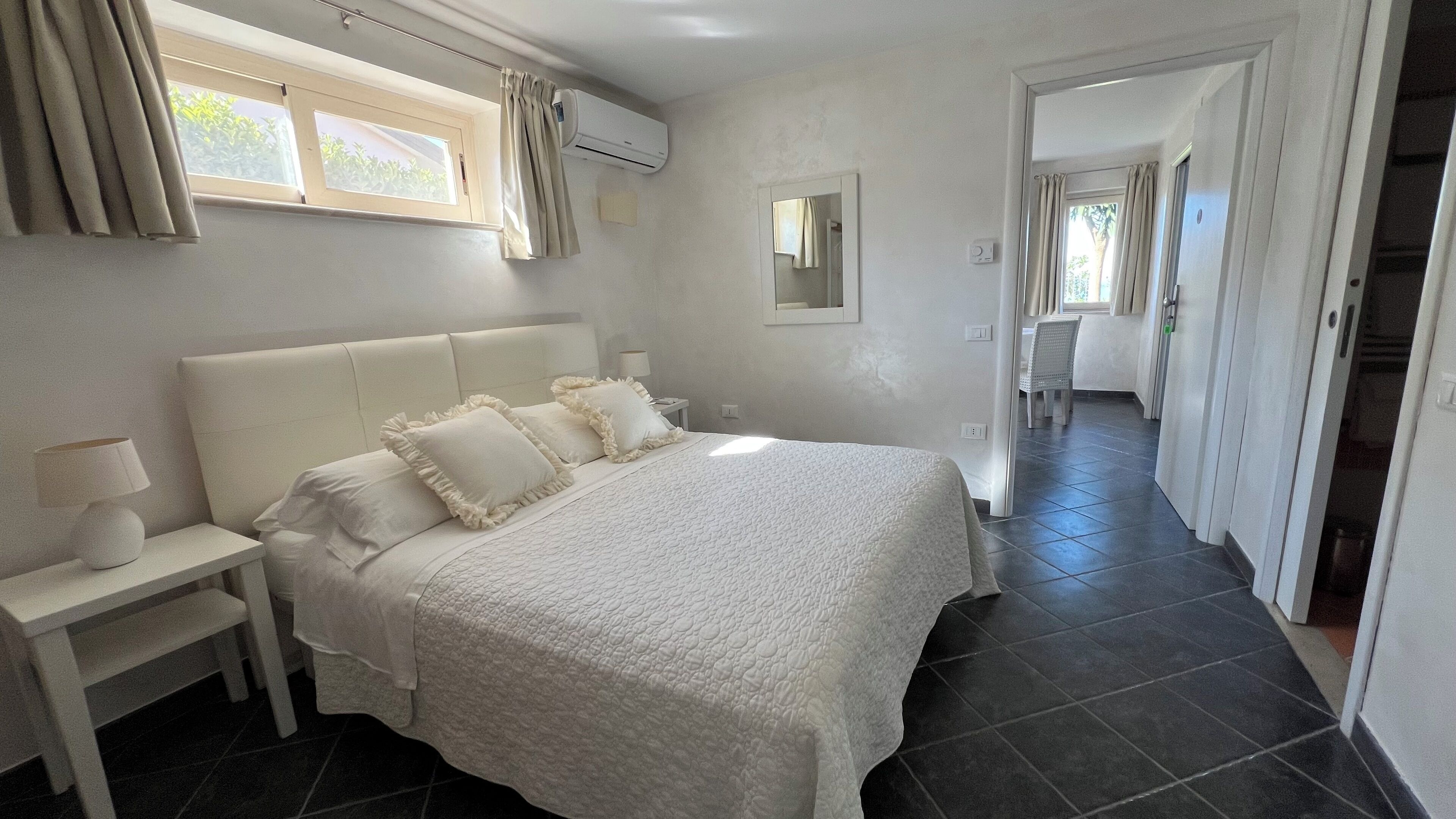 Un dormitorio con una cama grande, una mesita de noche, una ventana con cortinas, un equipo de aire acondicionado y un espejo en la pared.