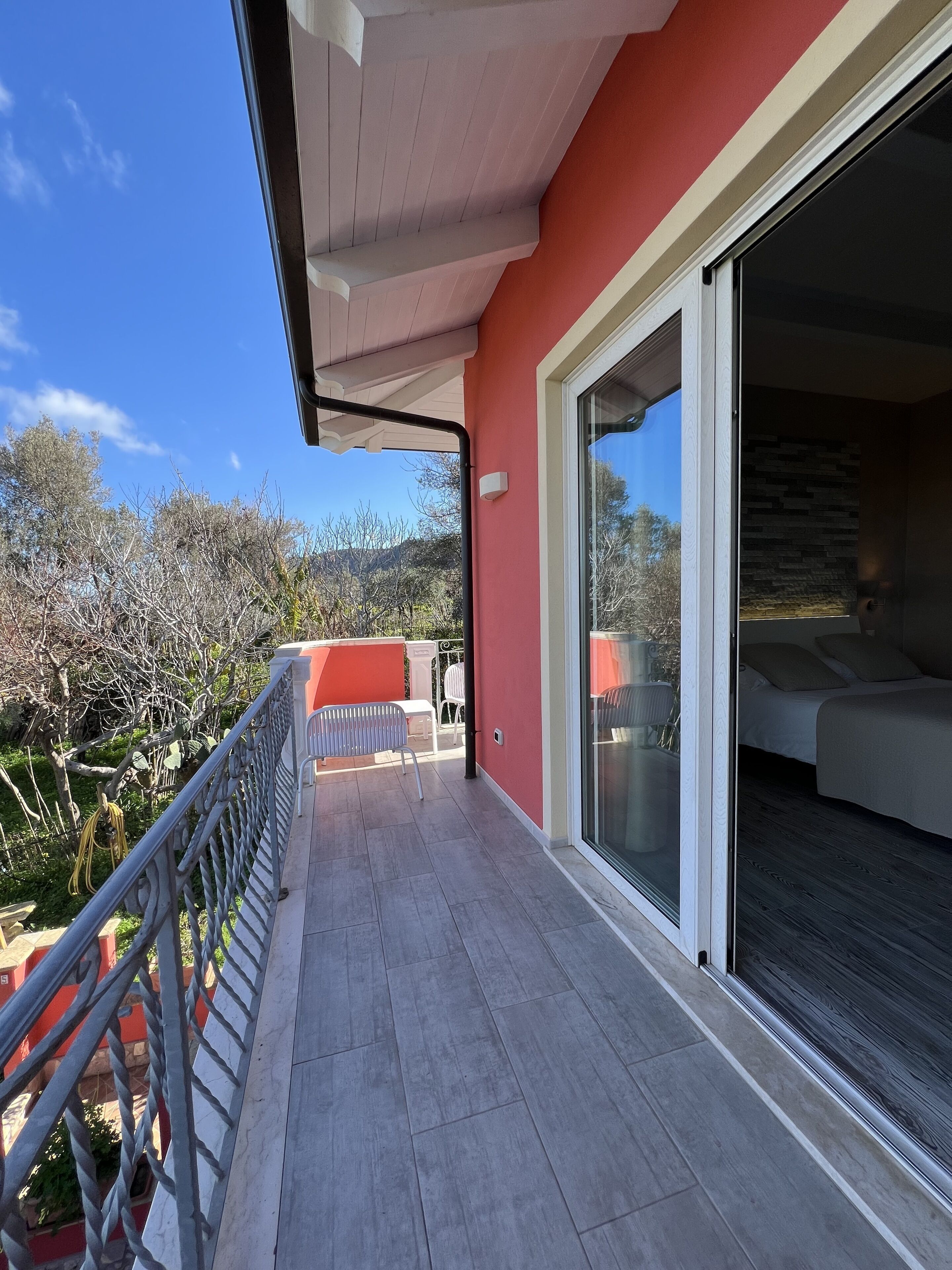 A balcony with a red wall, a sliding glass door leading to a bedroom, and a view of trees and hills.