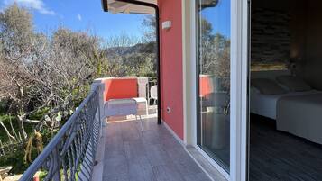 A balcony with a red wall, a sliding glass door leading to a bedroom, and a view of trees and hills.