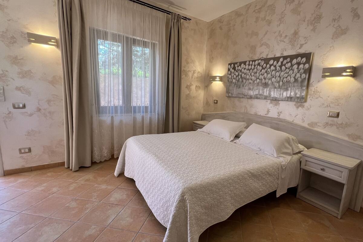 A bedroom with a large bed, two bedside tables, a nightstand, a painting on the wall, and a window with curtains.
