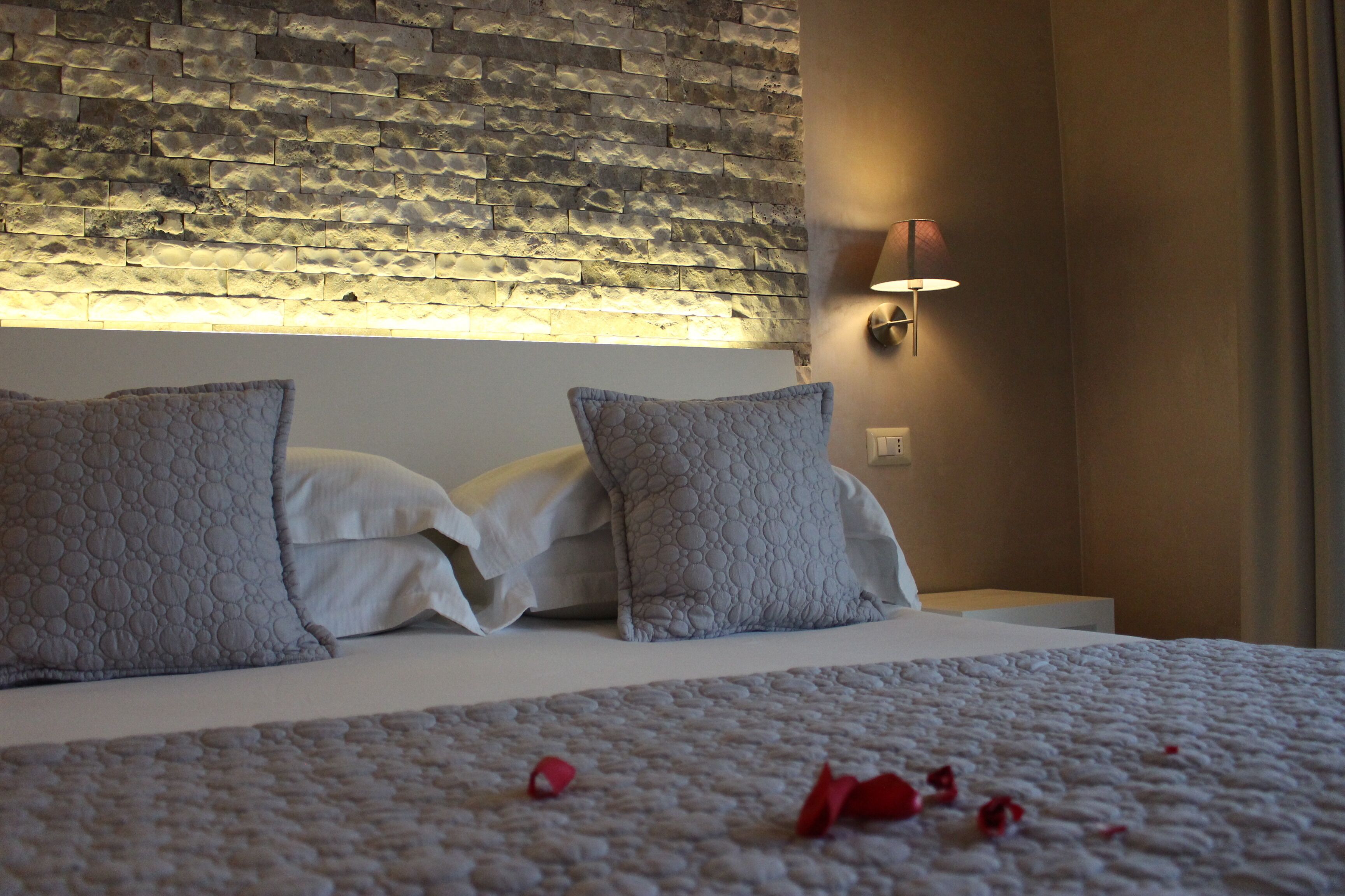 Una cama bien hecha con una manta texturizada y dos almohadas, una pared de piedra con una lámpara sobre ella, y una lámpara de pared a la derecha.