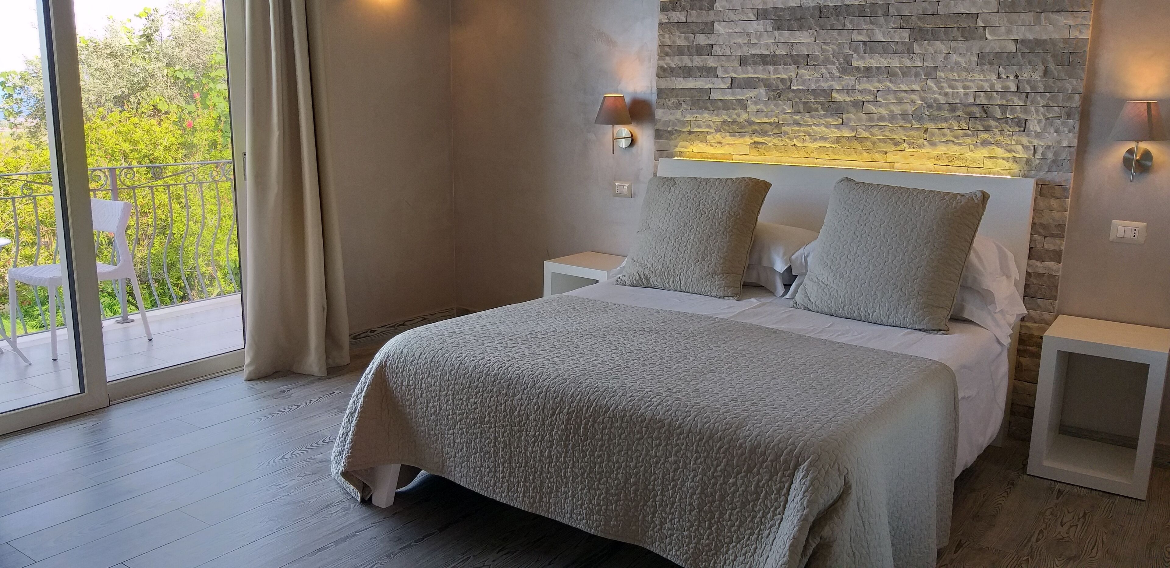 A modern bedroom with a large bed, a stone wall, and a balcony with outdoor furniture.