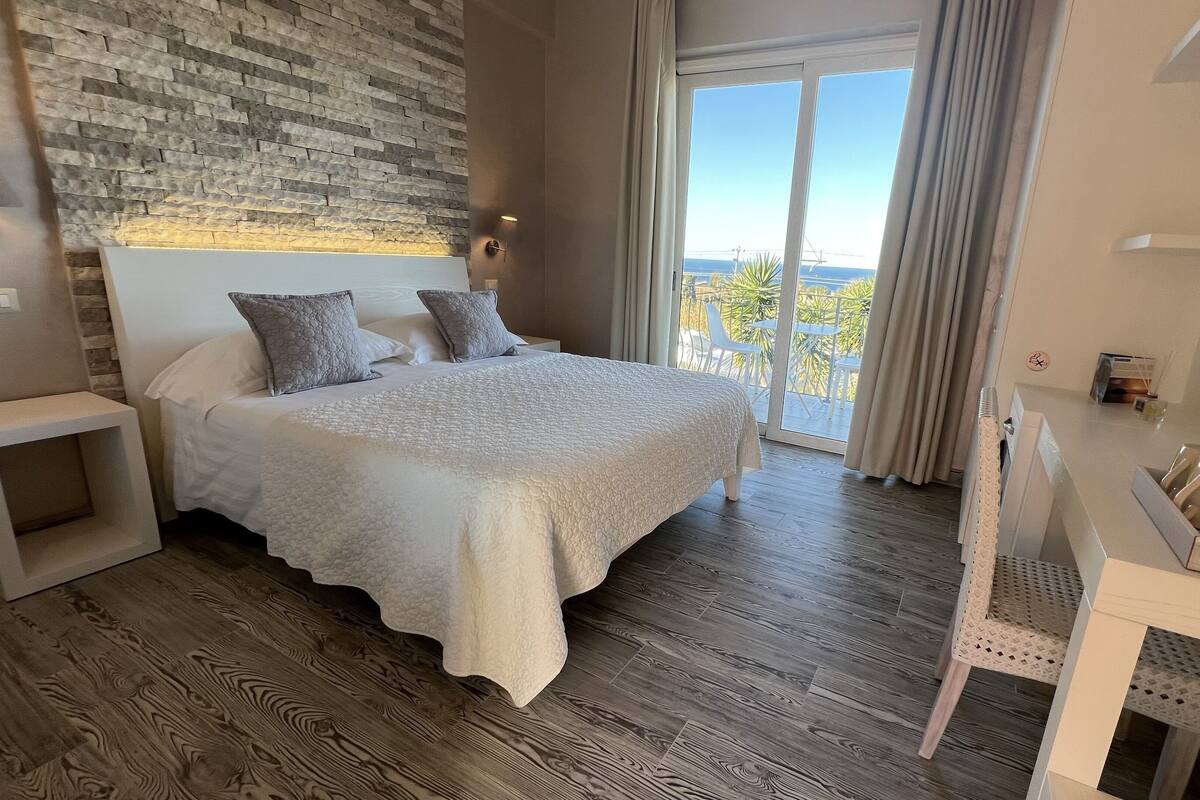 A bedroom with a large bed, a wooden floor, a stone wall, and a view through a glass door.