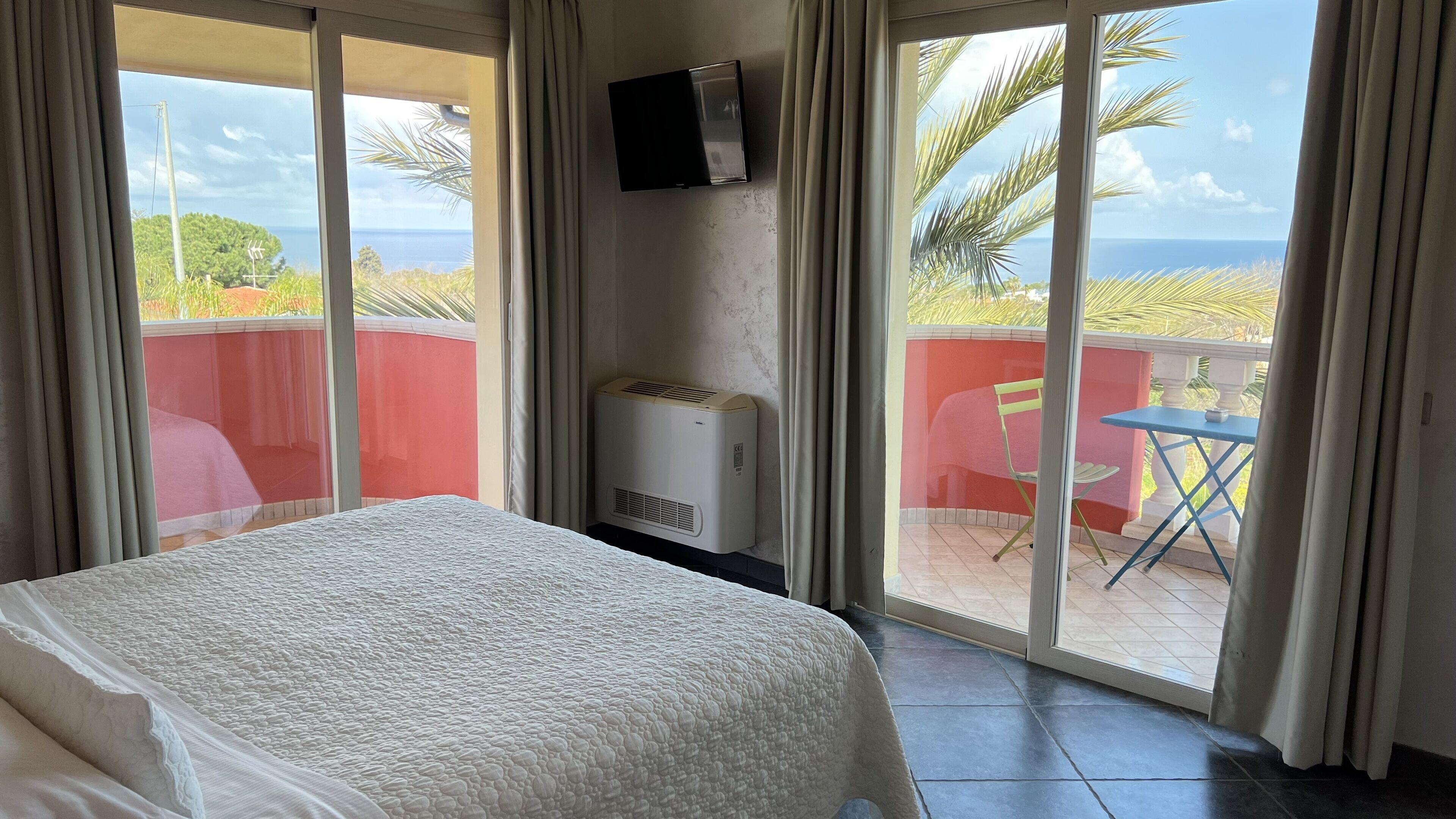 Une chambre d’hôtel avec un lit, un téléviseur fixé au mur, une petite table et une chaise, et une vue sur l’océan grâce à de grandes portes coulissantes.