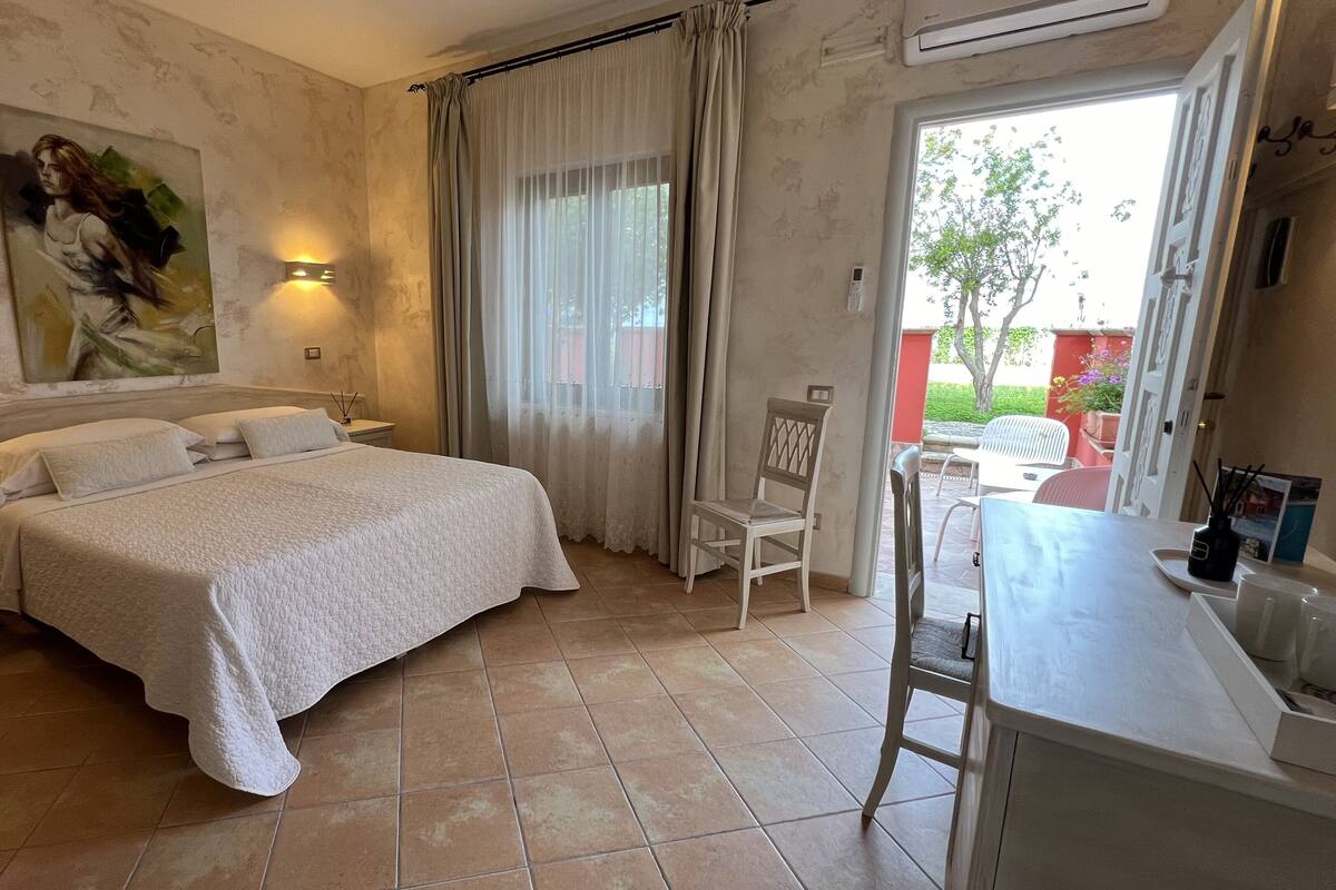 A hotel room with a large bed, a desk with a chair, a painting on the wall, and a view of the outside.