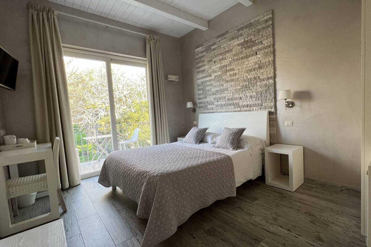 A bedroom with a large bed, a bedside table, a TV mounted on the wall, and a view of the outdoors.