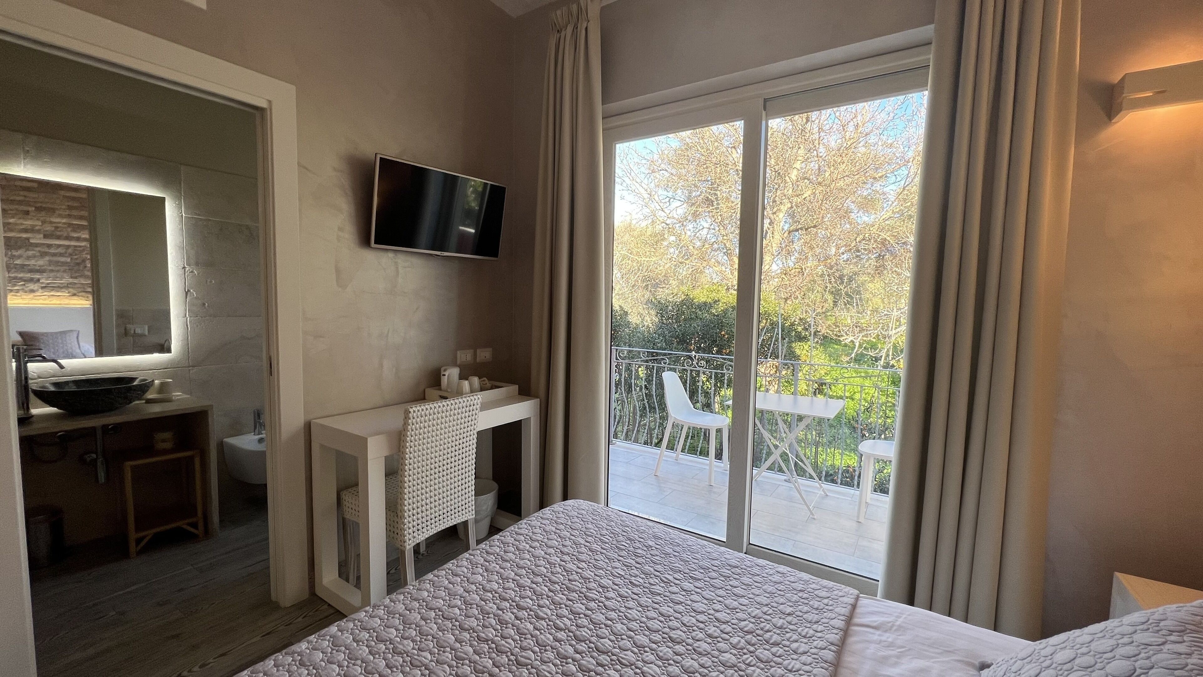 Ein Schlafzimmer mit einem großen Bett, einem an der Wand befestigten Fernseher, einem Badezimmer mit Waschbecken und Spiegel sowie einem Balkon mit Blick auf Bäume.