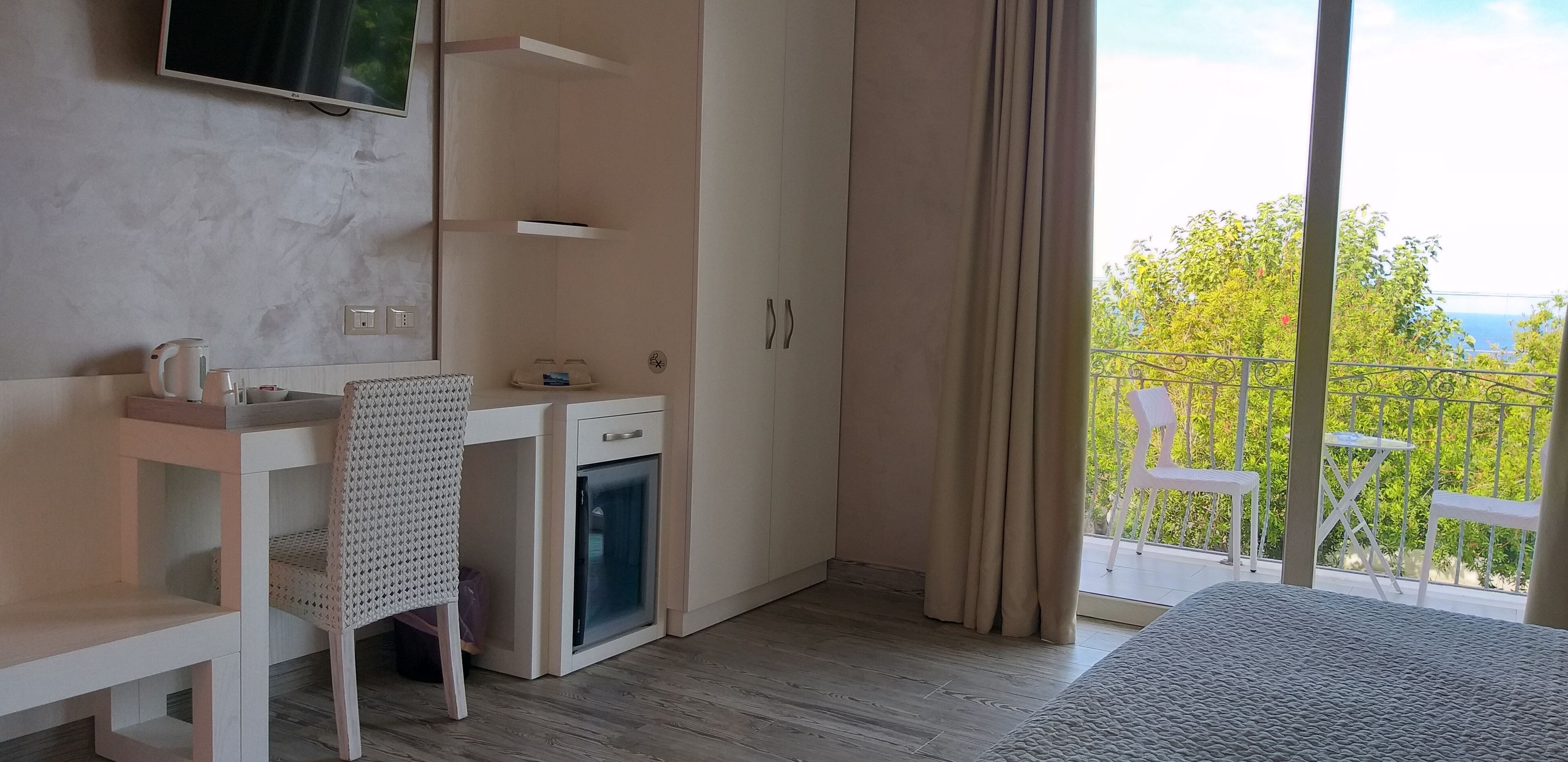 A bedroom with a bed, a desk, a chair, a wardrobe, a small fridge, and a balcony with a view of greenery and the sky.