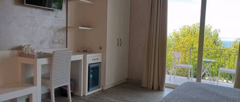 Un dormitorio con cama, escritorio, silla, armario, refrigerador pequeño y balcón con vista a la vegetación y el cielo.