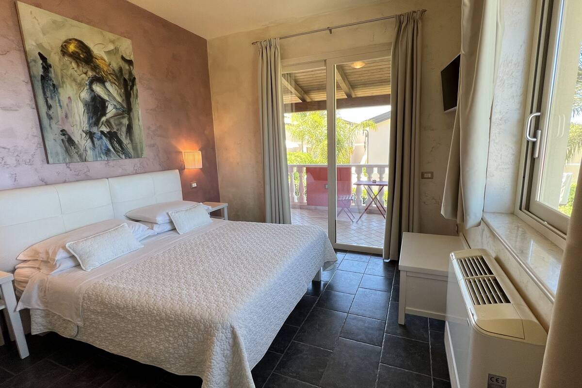 A bedroom with a bed, a nightstand, a wall-mounted lamp, a window with curtains, and a view of the outside balcony.