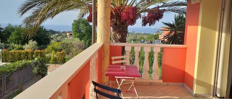 A balcony with a red table and bench, overlooking a garden and the sea.