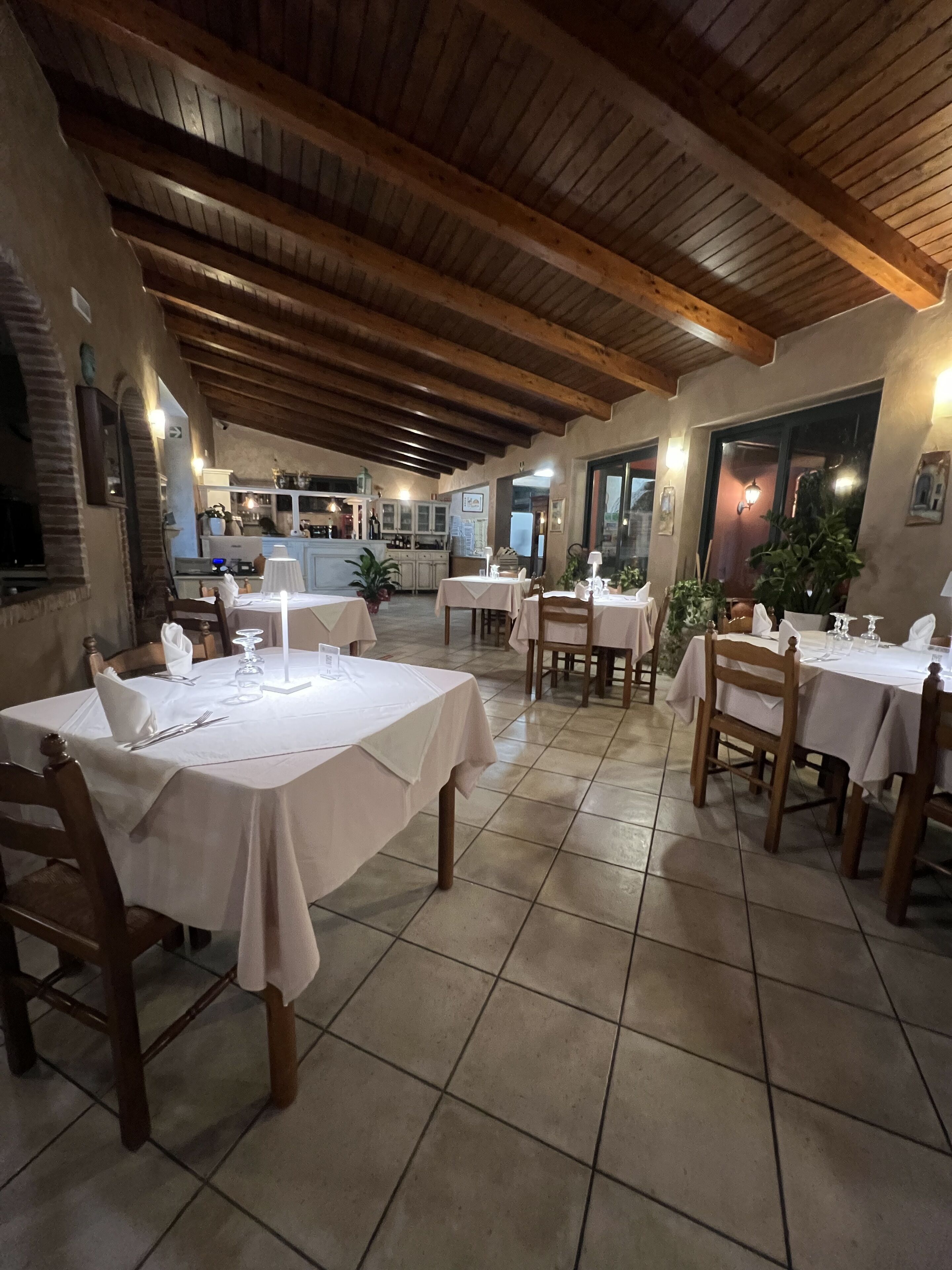 A restaurant with wooden beams on the ceiling, tiled floors, and tables set with white tablecloths and chairs.