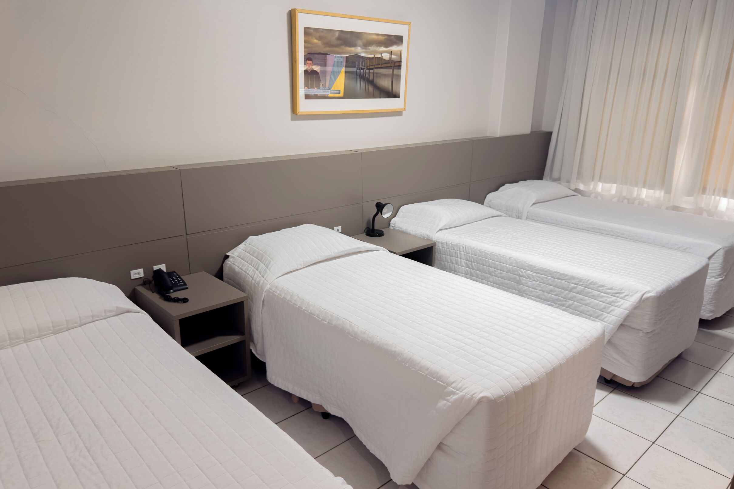 A hotel room with three beds, a nightstand with a phone, a framed picture on the wall, and a window with curtains.