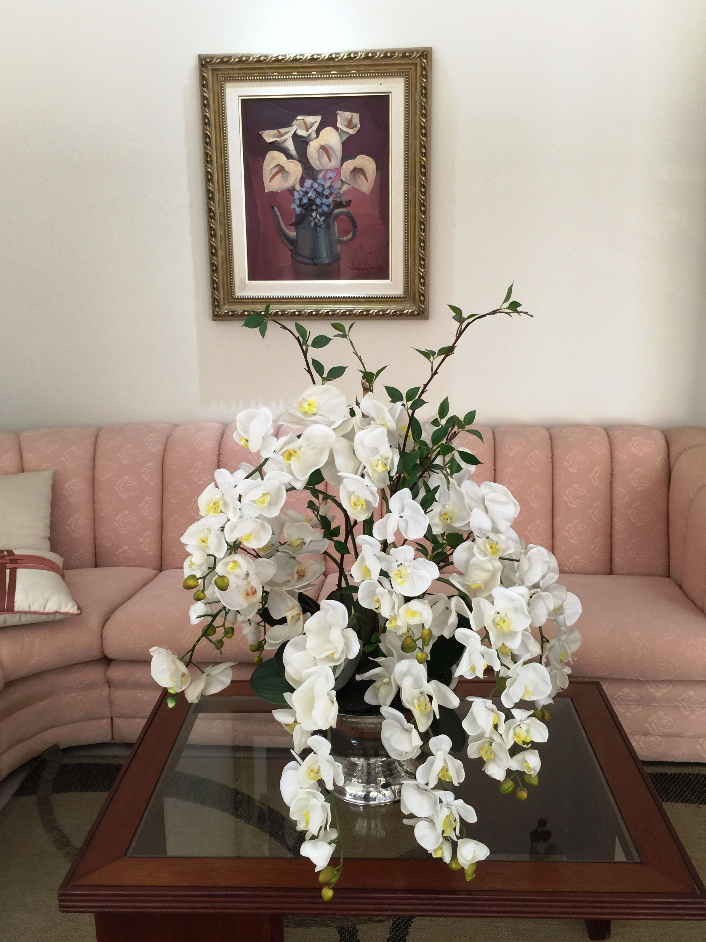 A living room with a glass coffee table in the center, a pink sofa with a cushion, and a framed painting on the wall.