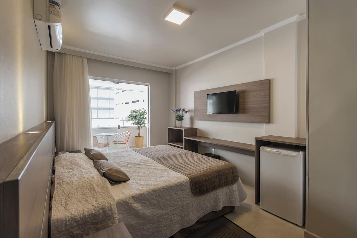 A hotel room with a bed, a TV mounted on the wall, a small fridge, and a window with curtains.