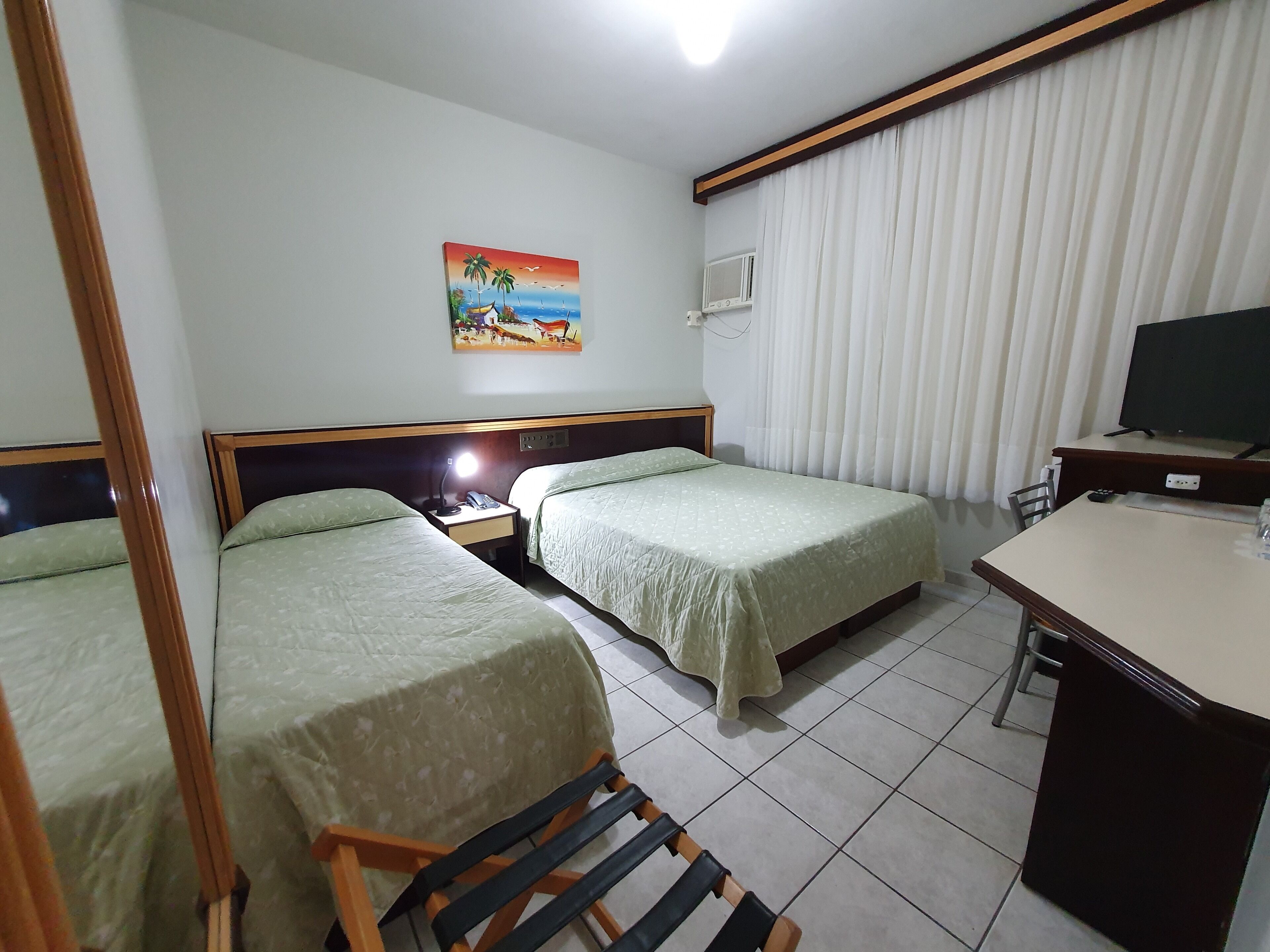 Habitación de hotel con dos camas individuales, un escritorio con televisor, un cuadro en la pared y una ventana con cortinas.
