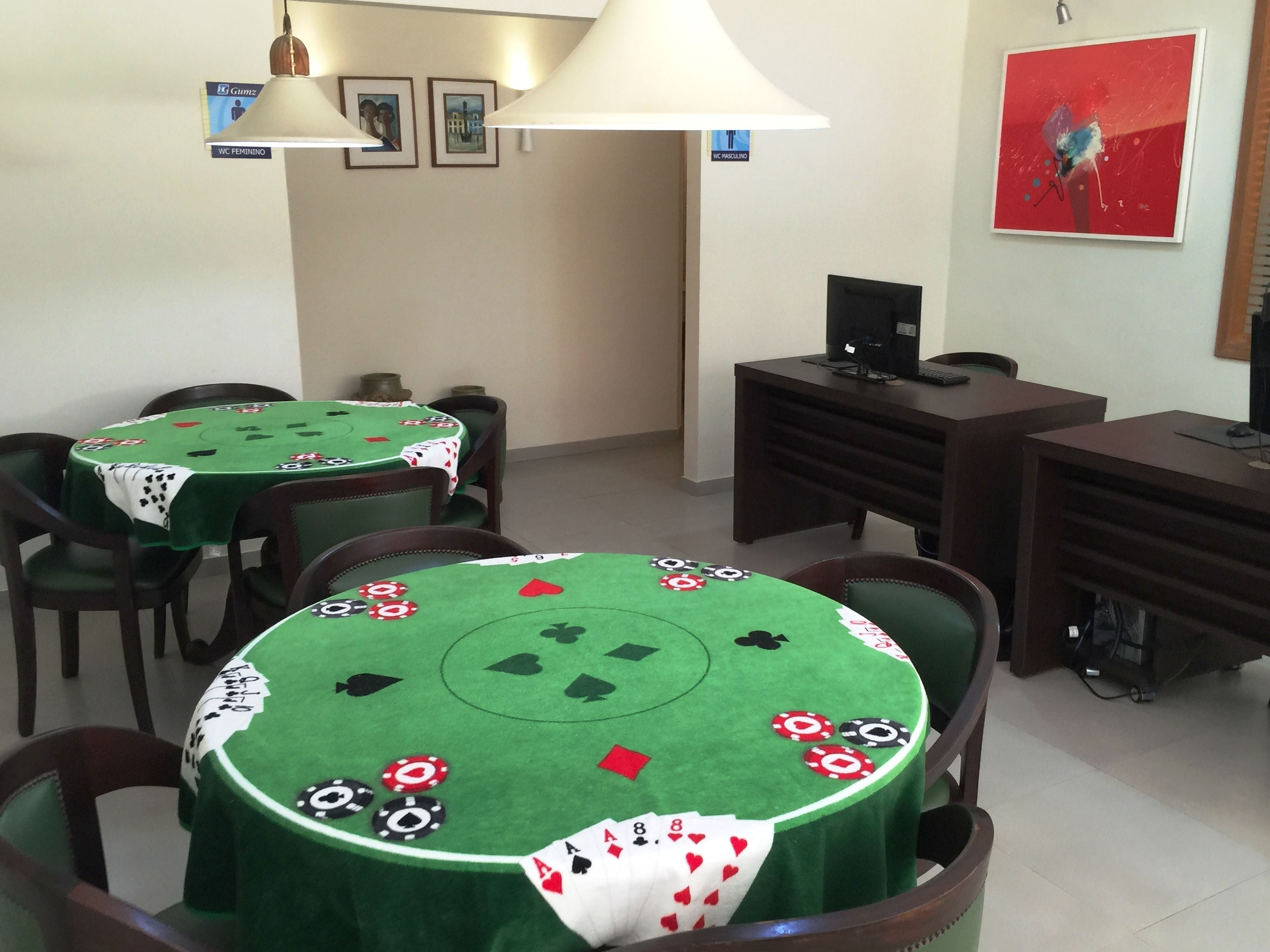 Ein Raum mit zwei mit grünem Filz bezogenen Tischen, an jedem Tisch Pokerchips und Spielkarten. Um die Tische herum befinden sich Stühle, ein Schreibtisch mit einem Computer und eine Lampe.