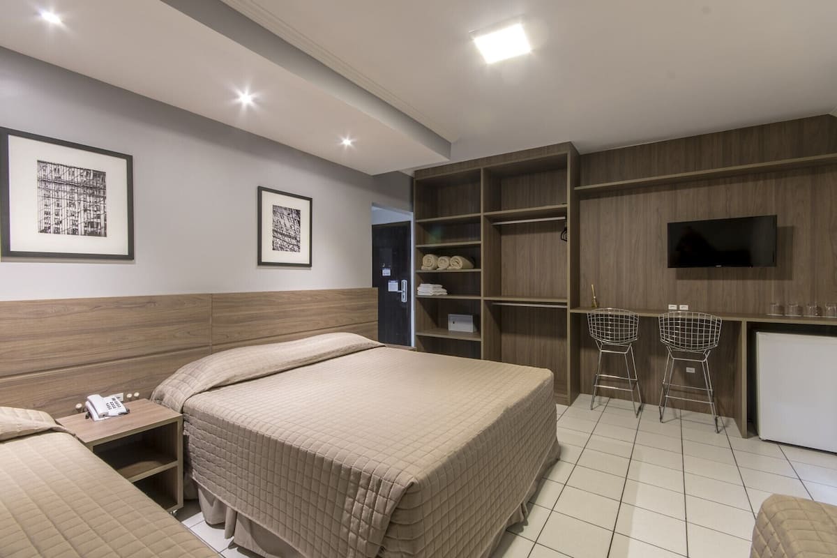 A hotel room with a bed, a bedside table with a phone, a TV, and a kitchenette area with two bar stools.