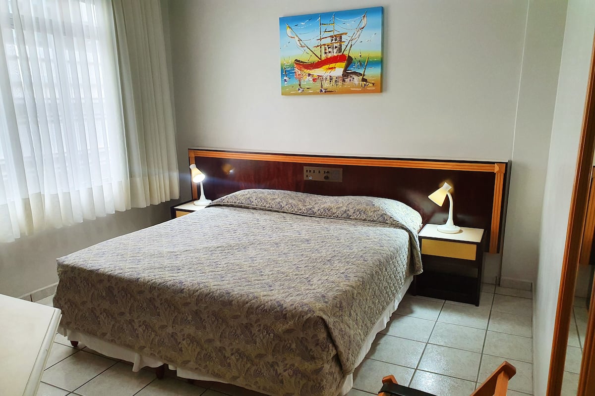 A neatly made bed with a patterned bedspread, two bedside lamps, a wooden headboard, and a painting of a ship on the wall.