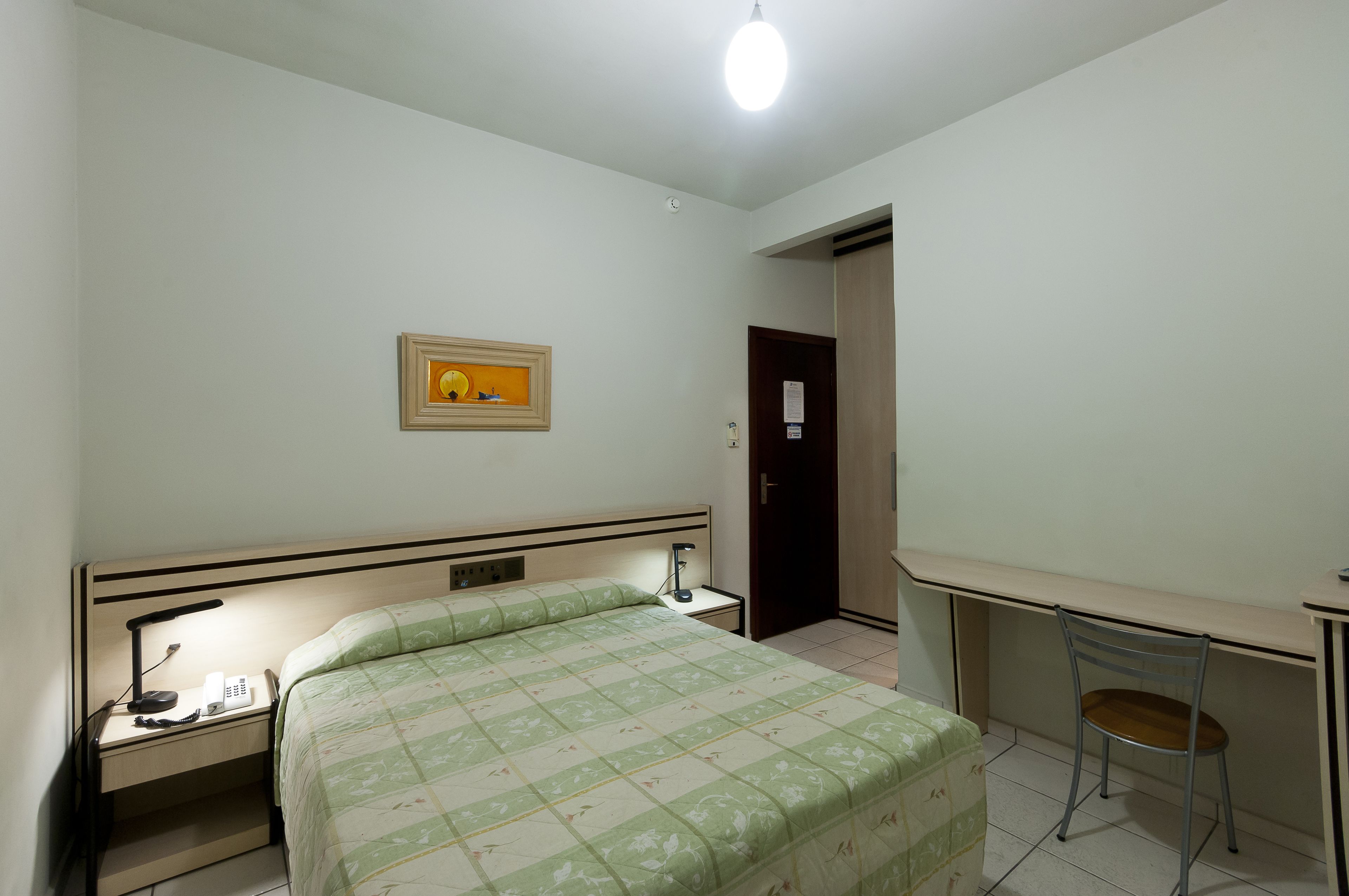 Un dormitorio con cama, mesita de noche, lámpara, escritorio, silla y una puerta.
