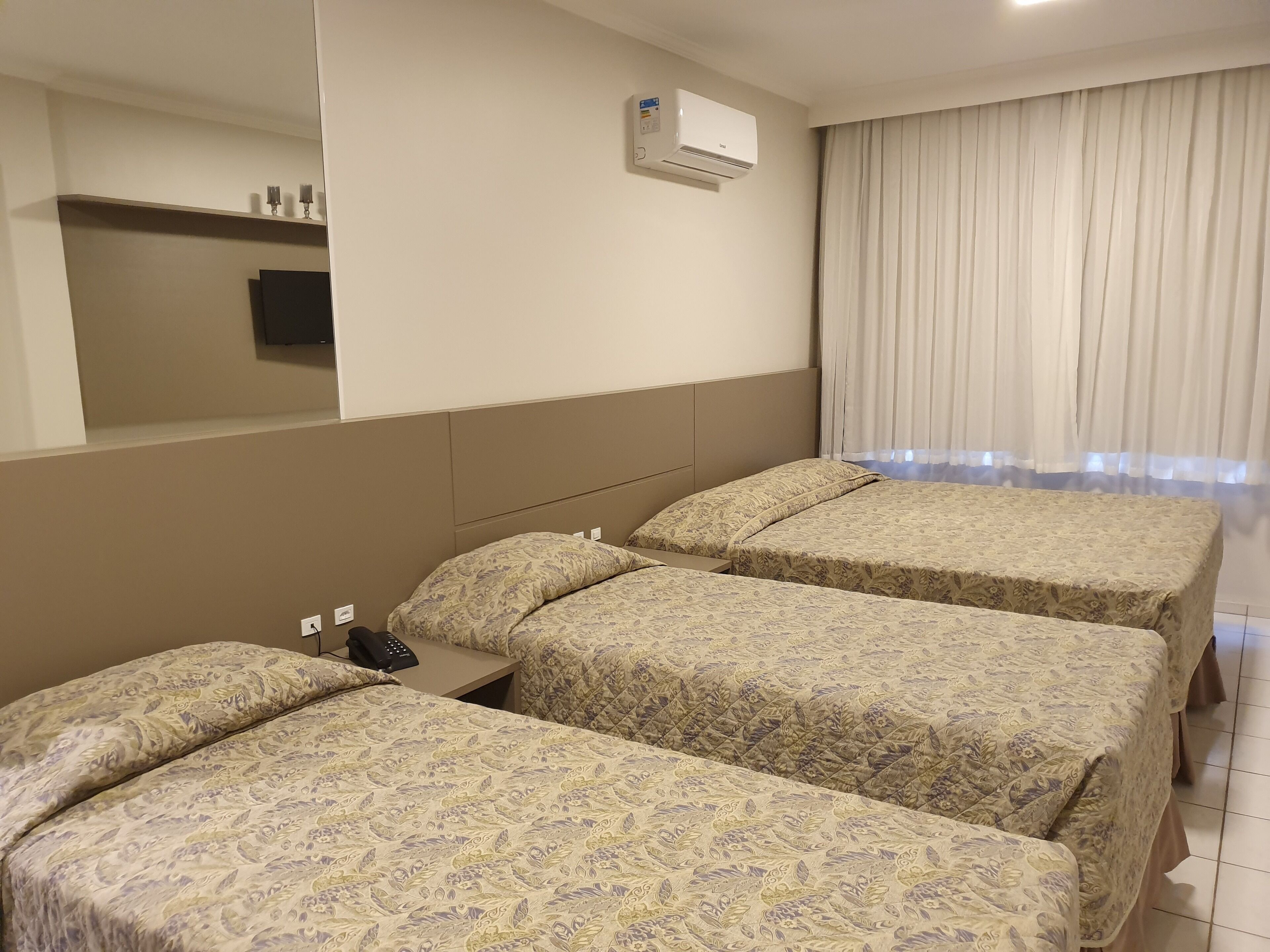 A hotel room with three beds, a bedside table with a phone, an air conditioning unit, and a window with curtains.
