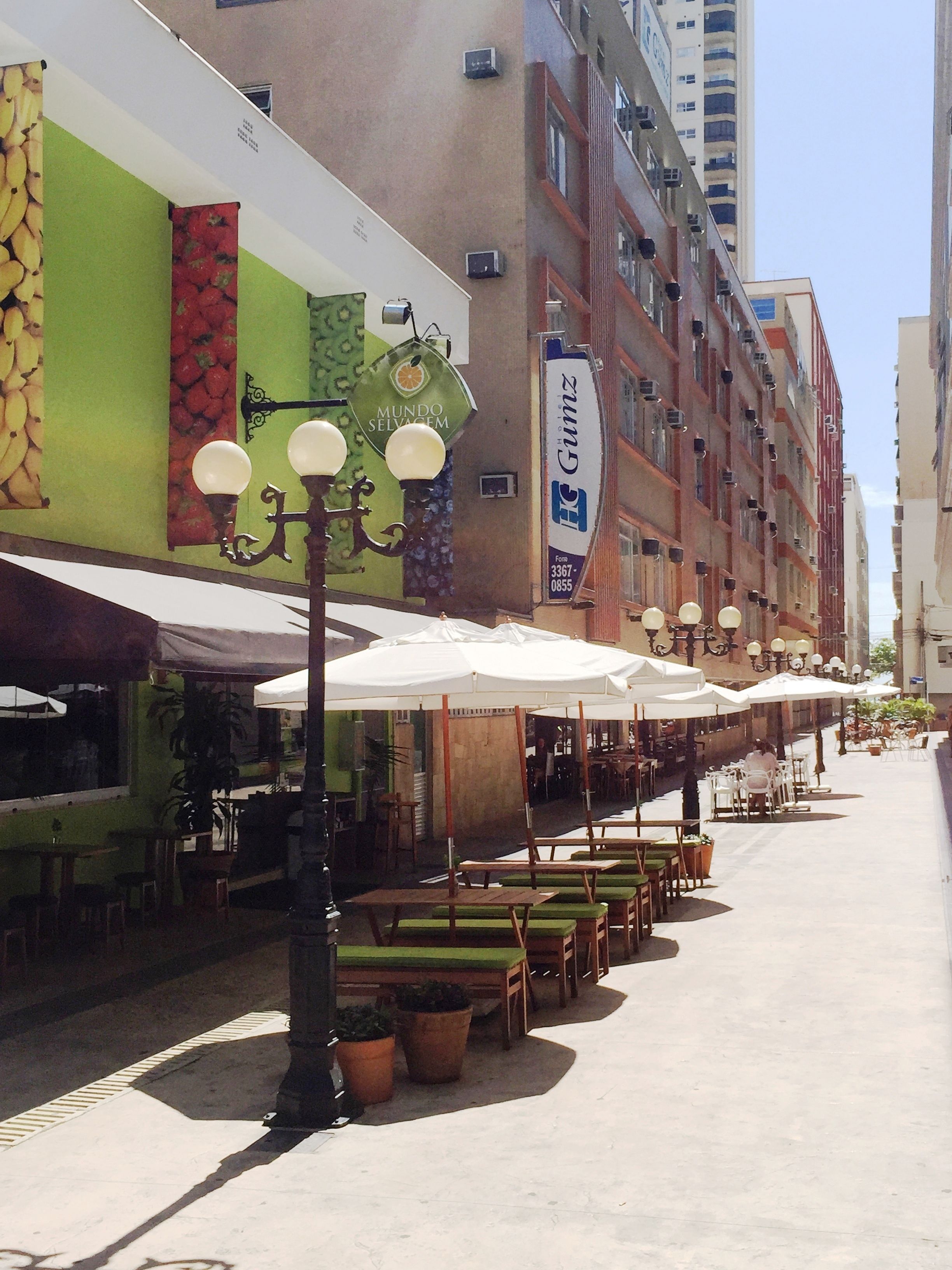 Un café al aire libre con mesas y sillas bajo sombrillas blancas, un farol y un edificio con un mural de frutas.