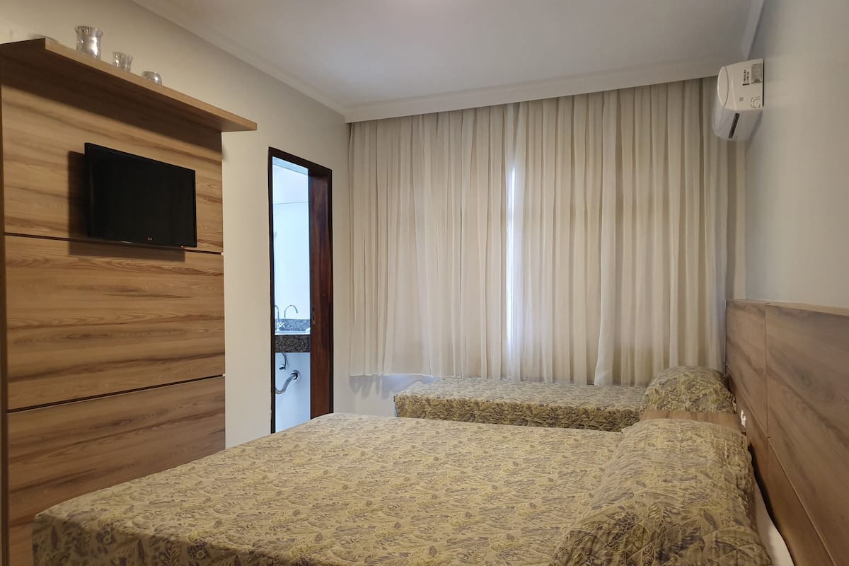 A hotel room with two beds, a TV on the wall, a wooden wardrobe, and a window with curtains.