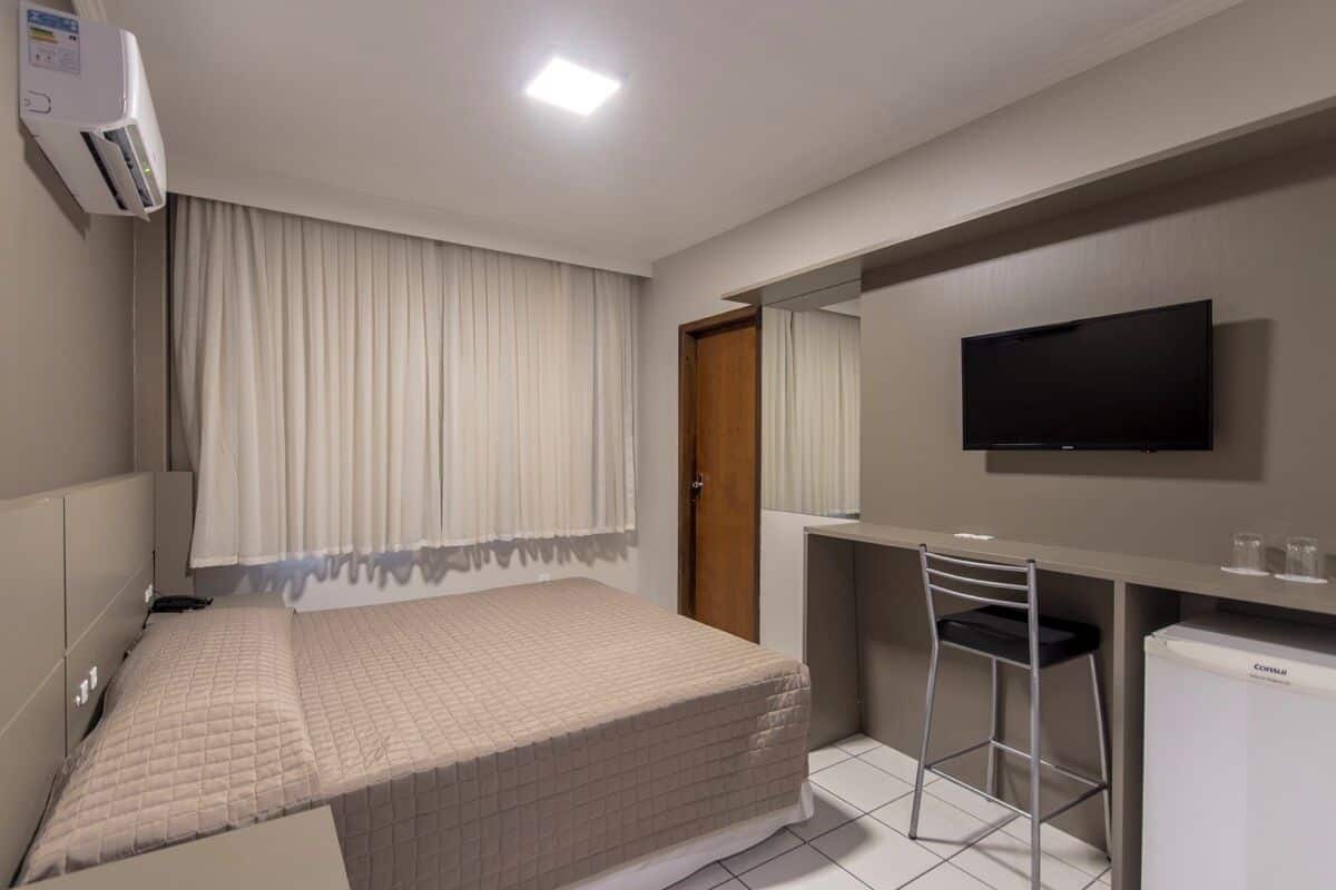 A hotel room with a bed, a desk, a chair, a television, and an air conditioning unit.