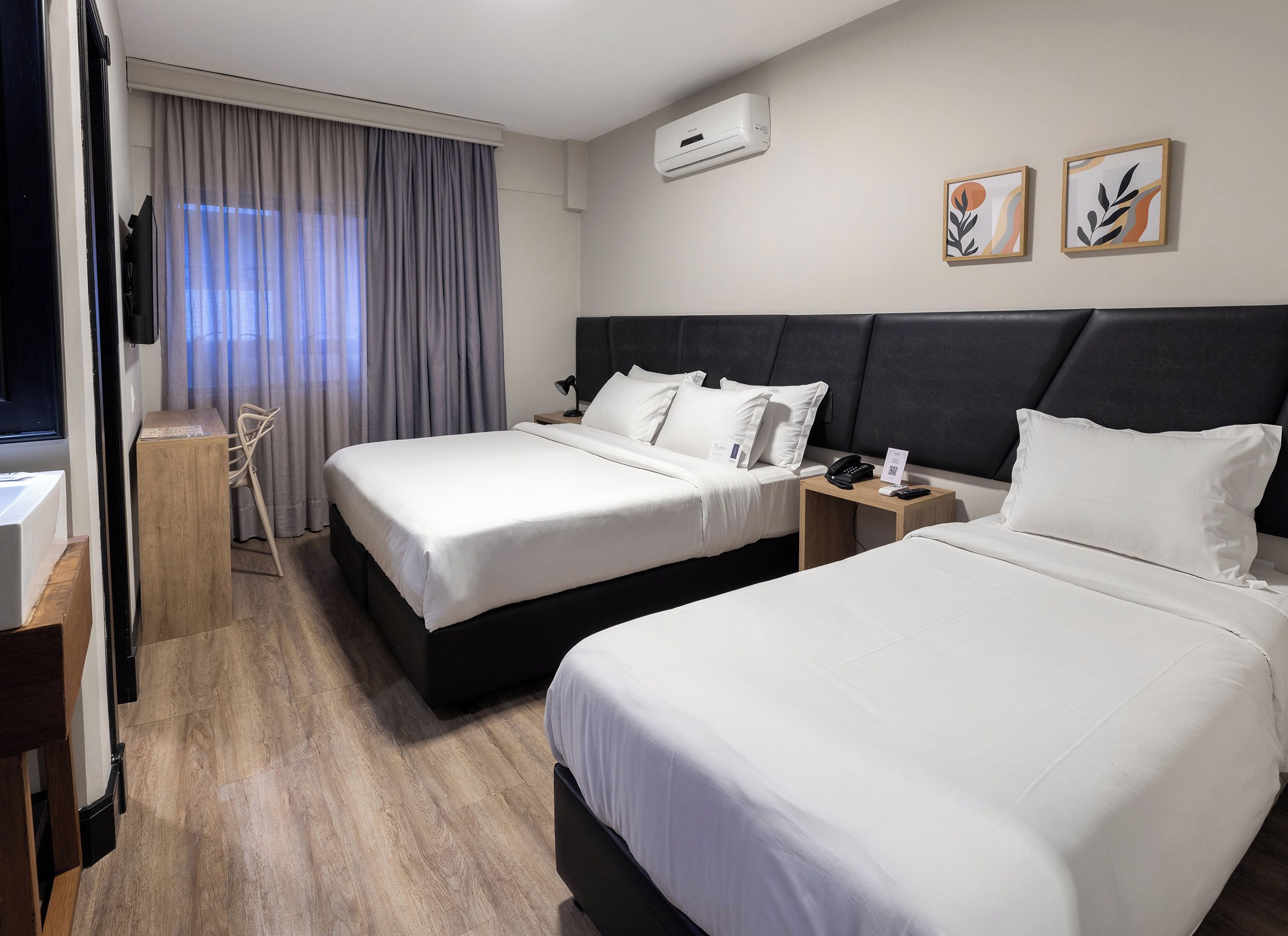 Quarto de hotel com duas camas, uma escrivaninha, uma cadeira, mesa de cabeceira com telefone e controle remoto, e ar condicionado na parede.