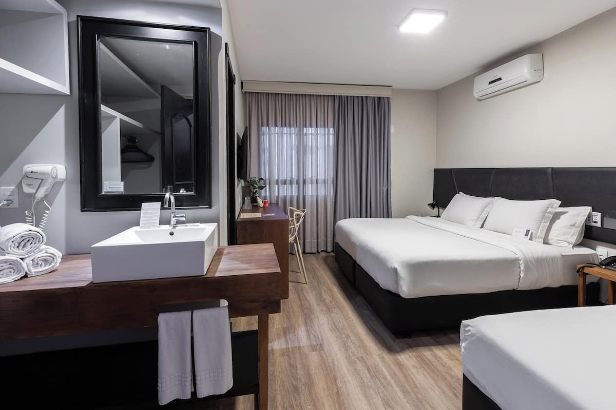 Une chambre d’hôtel avec un grand lit, une table de chevet, un bureau avec un miroir, un lavabo blanc et un sèche-cheveux.