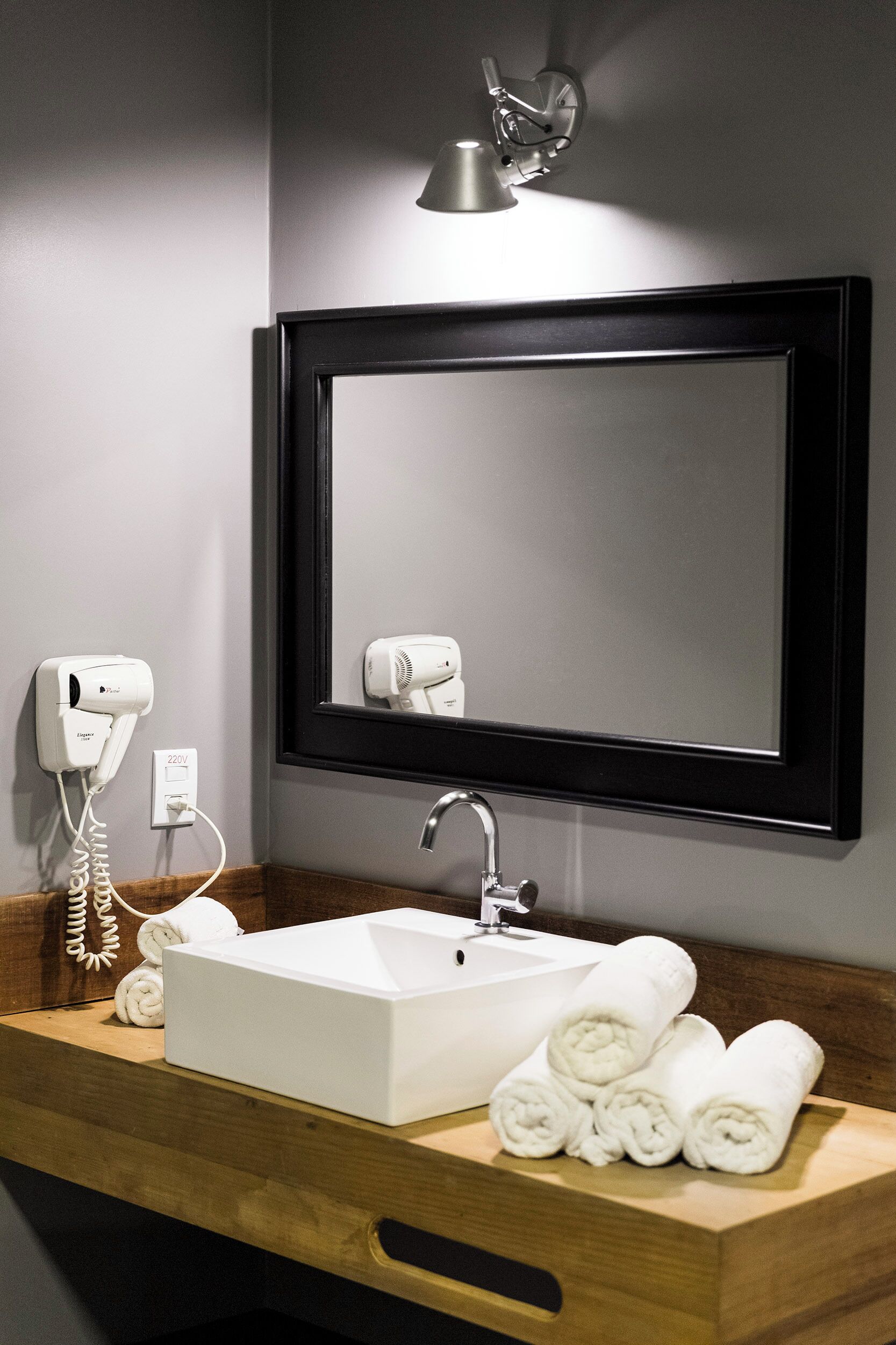 Une salle de bain avec un lavabo blanc, un miroir, un sèche-cheveux et des serviettes posées sur un meuble-lavabo en bois.