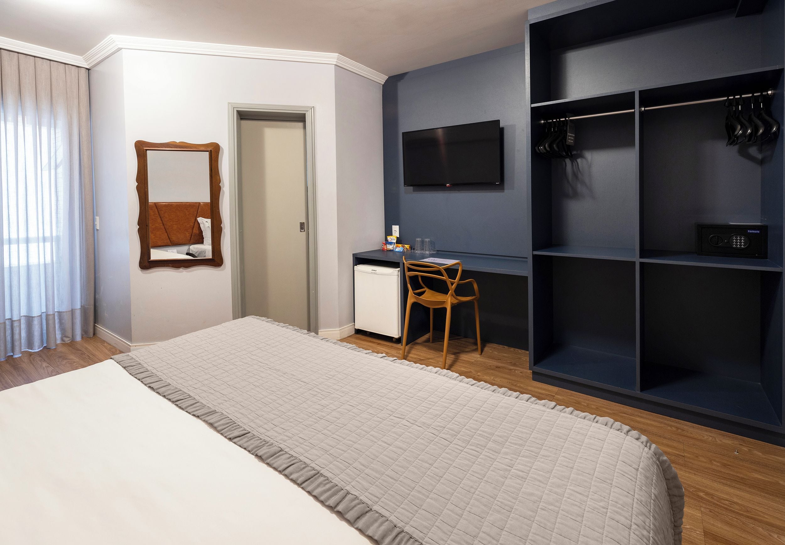 A hotel room with a bed, a desk, a chair, a wardrobe, a television, and a mirror.