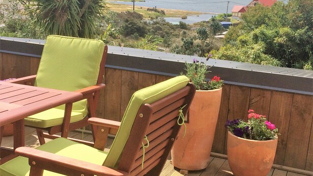 A wooden deck with two green cushioned chairs and a table, overlooking a lake with houses in the background. - Lakeside City Apartments (Rotorua)