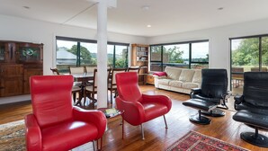A spacious living area with a wooden dining table, red and black leather chairs, a white sofa, and large windows offering a view of greenery. - Lakeside City Apartments (Rotorua)