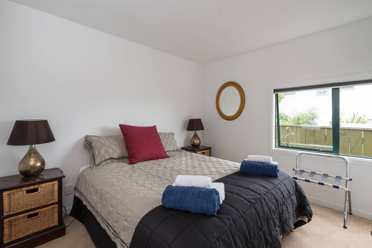 A bedroom with a bed, two bedside tables, a window, and a view of the outside.