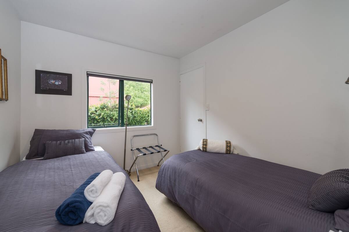 A bedroom with two beds, a window with a view of greenery, a framed picture on the wall, and a small rack near the window.