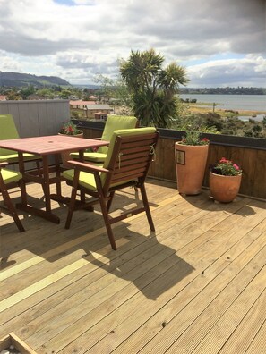 A wooden deck with a table and chairs, potted plants, and a view of a lake and hills. - Lakeside City Apartments (Rotorua)