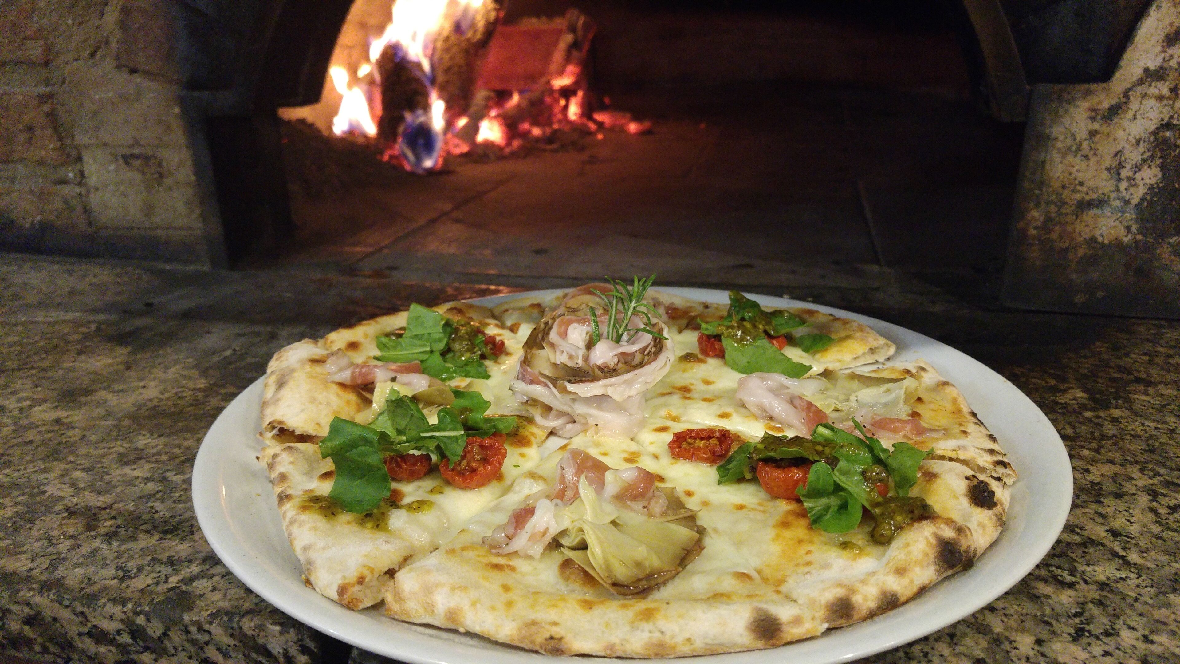 Pizza con diferentes ingredientes sobre un plato blanco, colocada frente a un horno de ladrillo con fuego encendido en su interior.