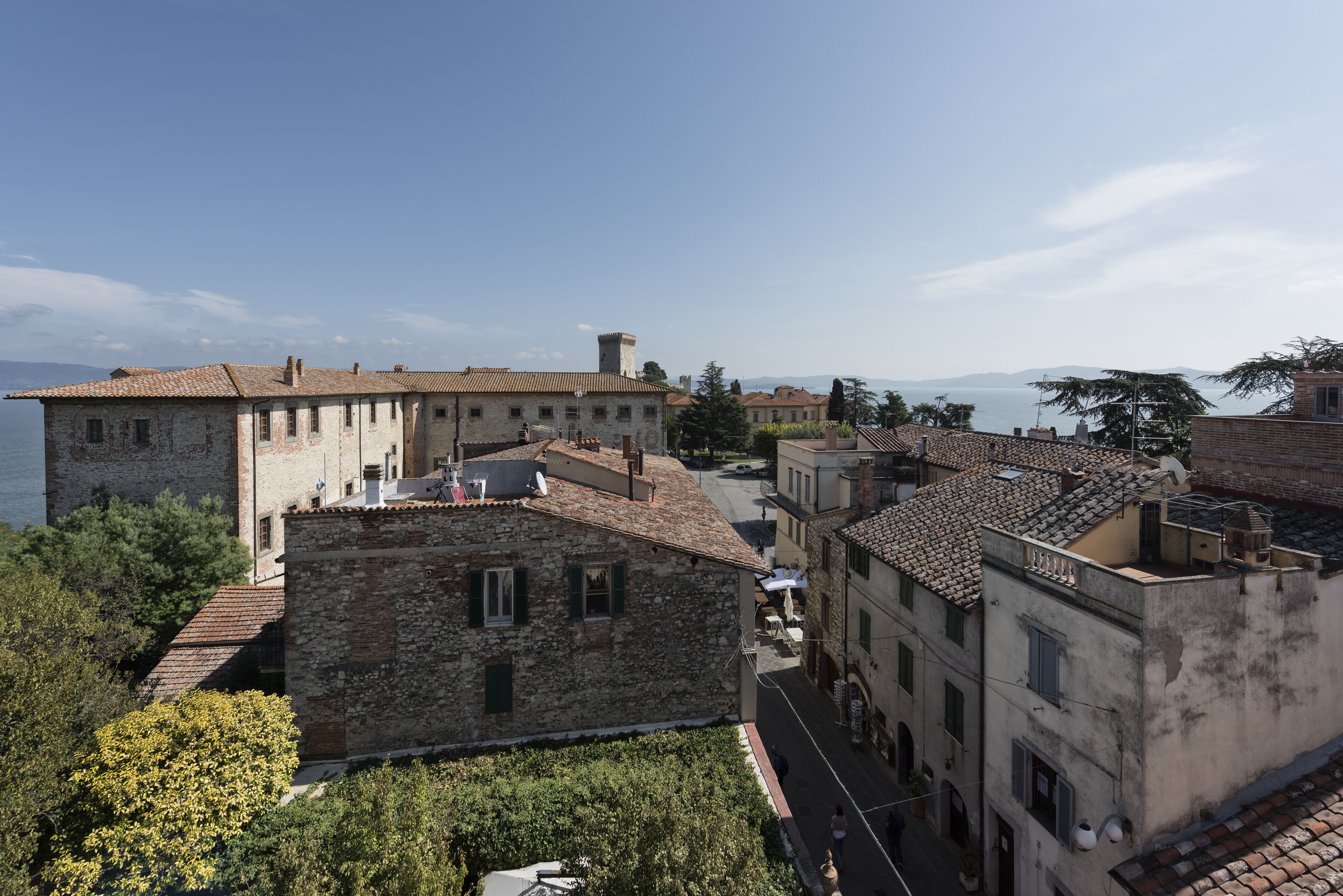 A historic stone building with a tiled roof, adjacent to a body of water and other buildings.