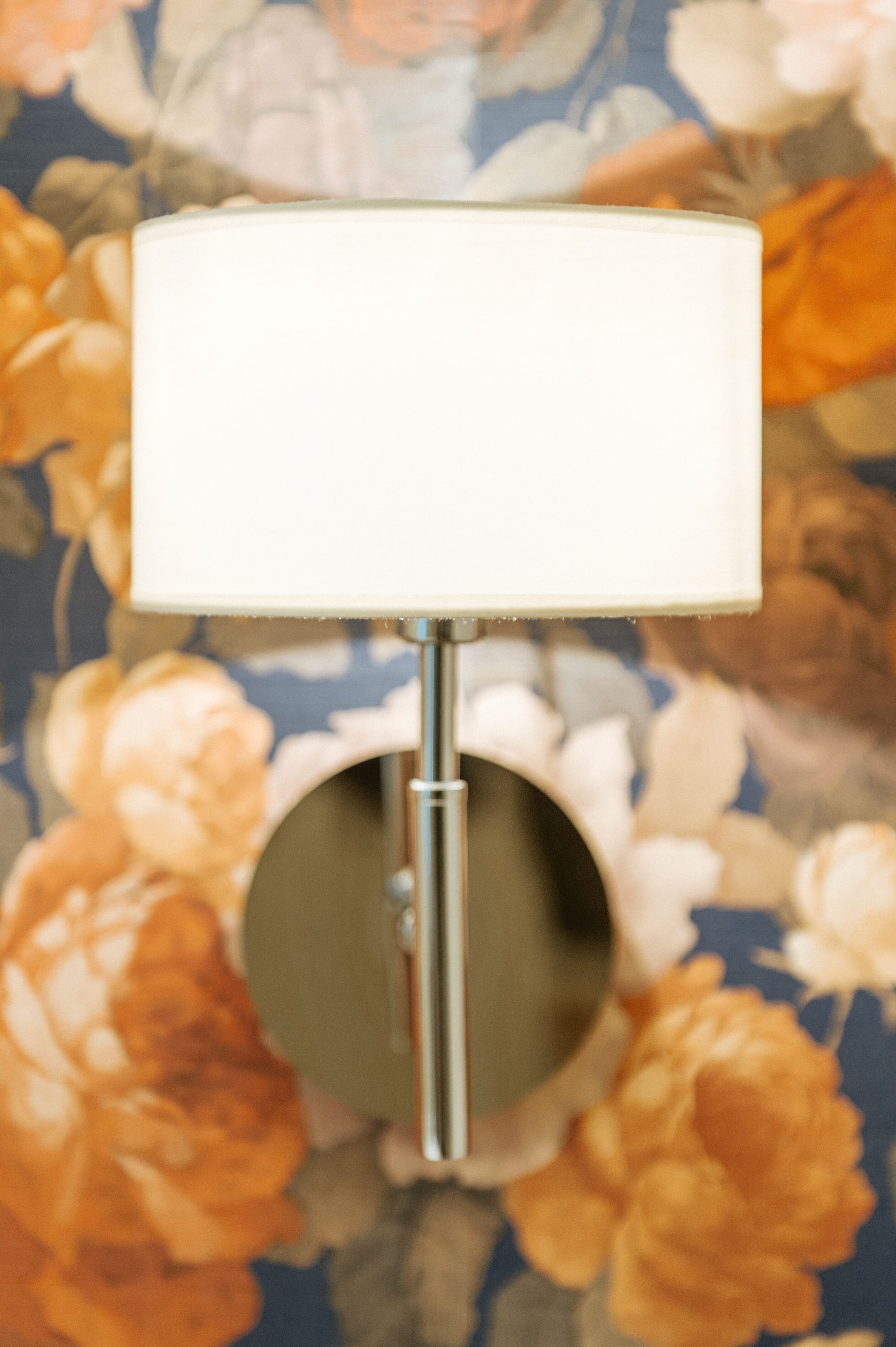 A wall-mounted lamp with a white shade and a metallic base, set against a floral wallpaper background.