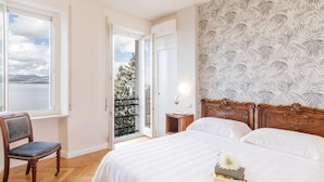 Une chambre avec un grand lit, une chaise, une table de chevet et une vue sur un arbre par une porte ouverte.