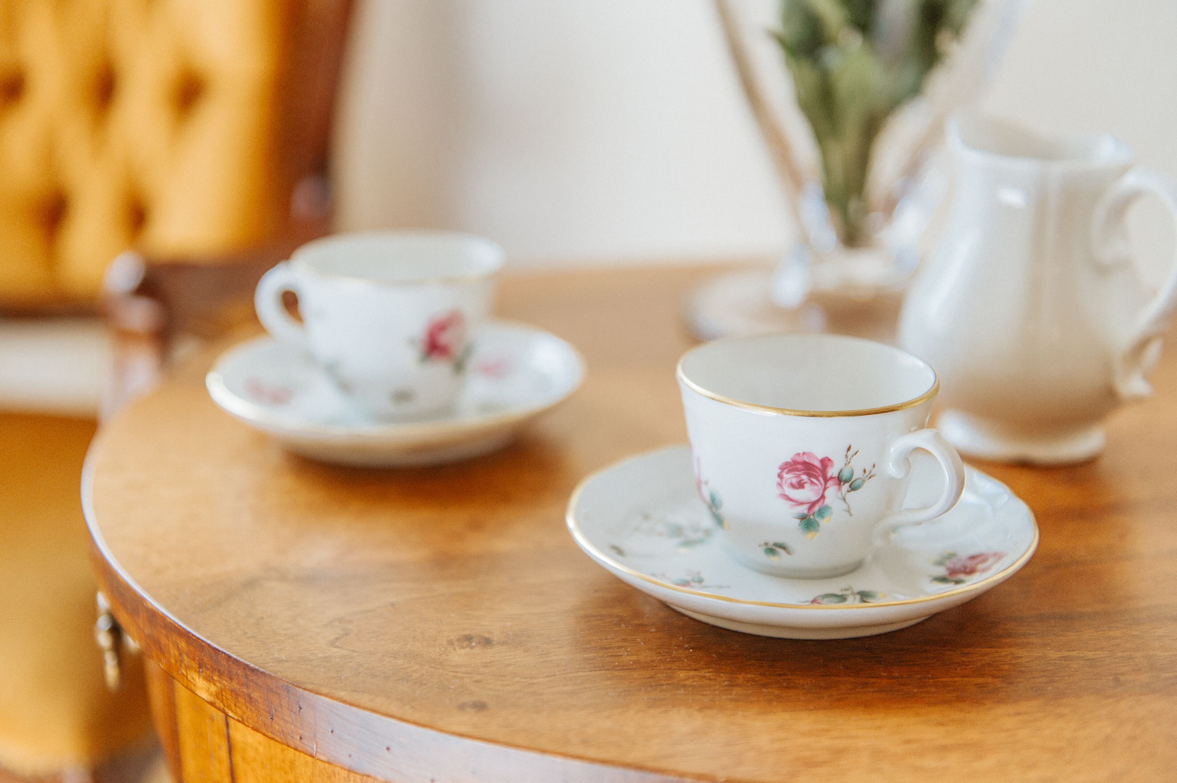 Une table ronde en bois, sur laquelle sont posées deux tasses blanches ornées de motifs floraux, ainsi qu’une théière assortie.