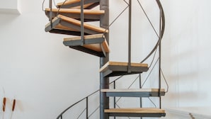 Un escalier en colimaçon avec des marches en bois et des rampes en métal, dans une pièce aux murs blancs et dotée d’un radiateur.