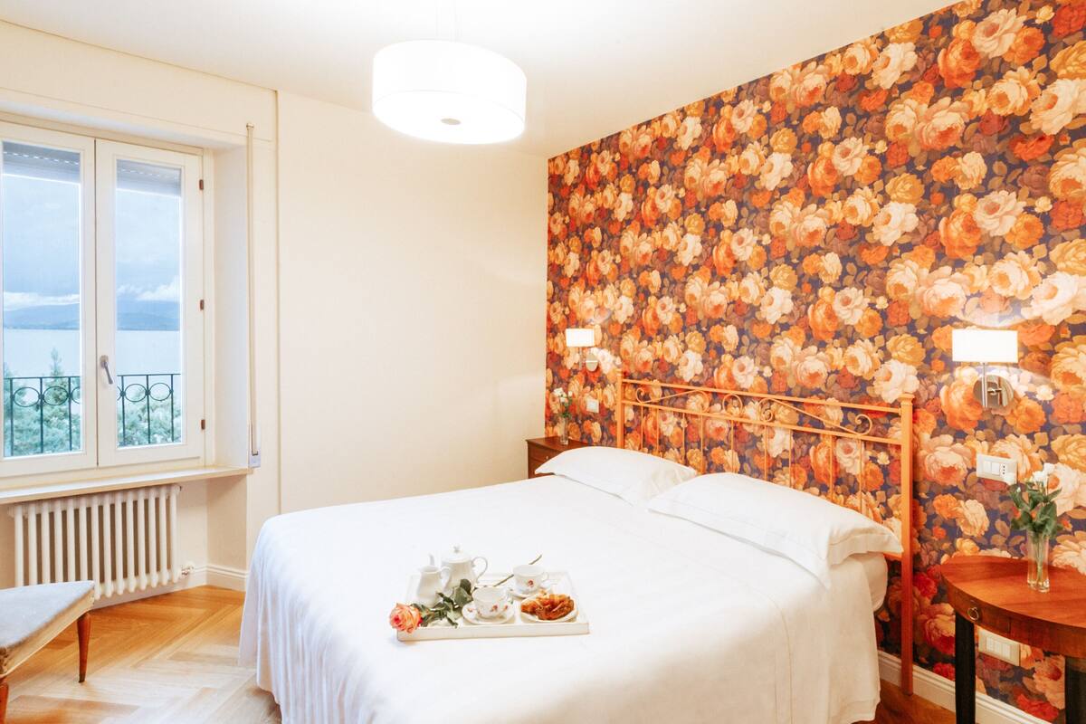 Une chambre avec un grand lit, une table de chevet, une fenêtre donnant sur un beau paysage et un papier peint à motifs floraux.