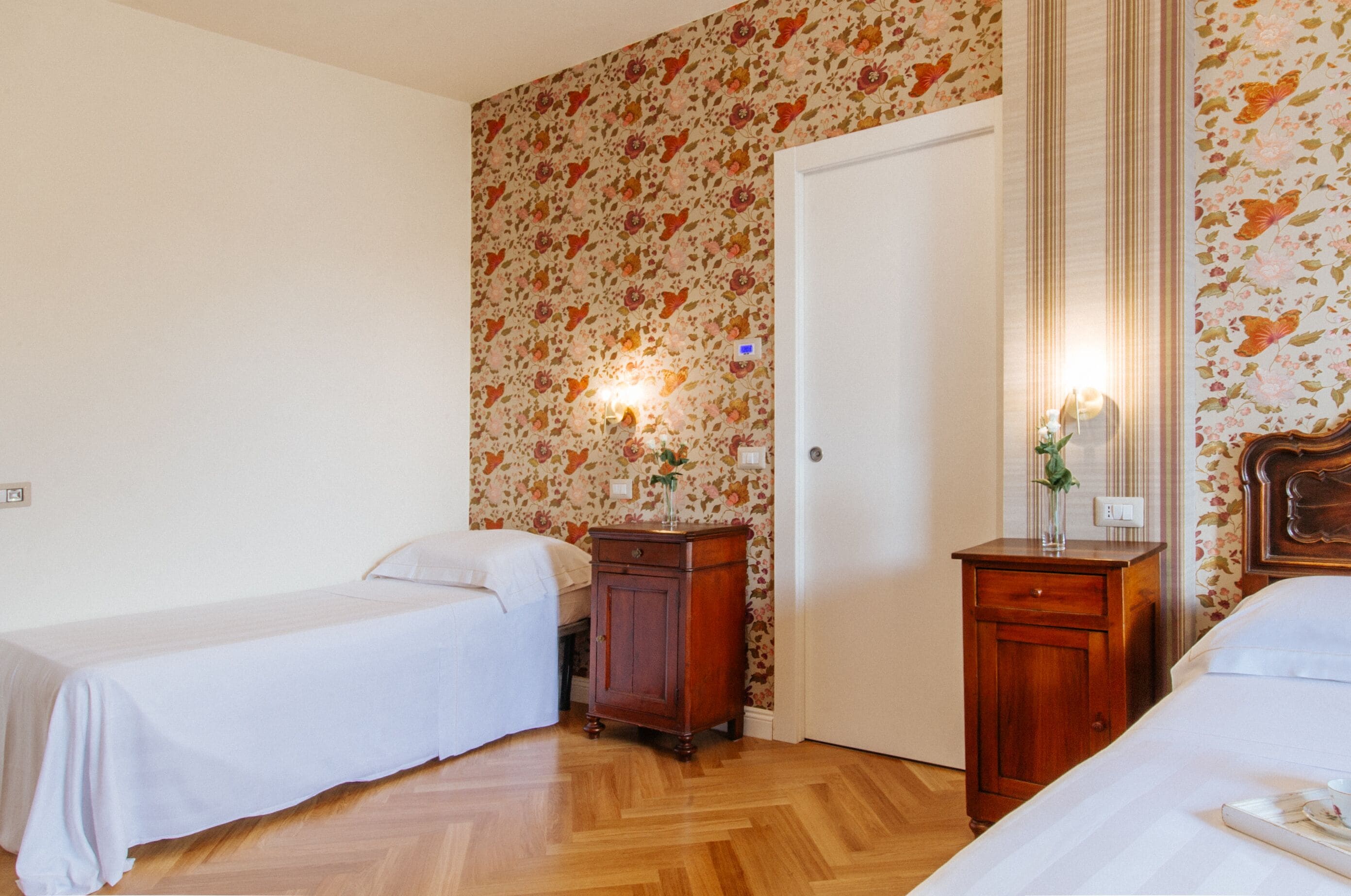 A room with two single beds, wooden flooring, a floral wallpaper, a door, and a nightstand with a lamp.