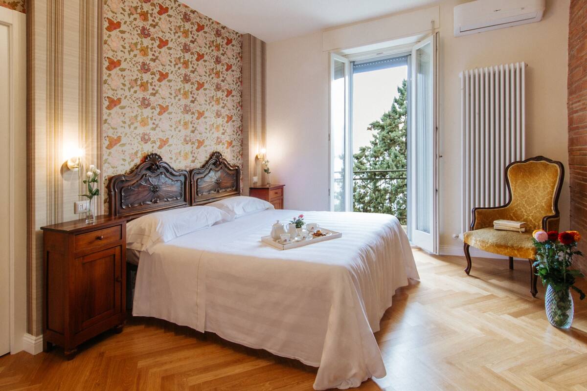 Une chambre avec un grand lit, des tables de chevet en bois, une chaise, un radiateur et une fenêtre donnant sur des arbres.