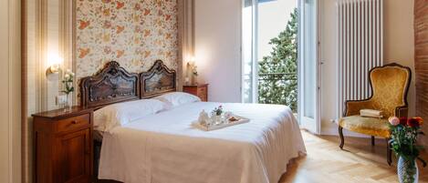 A bedroom with a large bed, wooden nightstands, a chair, a radiator, and a window with a view of trees.