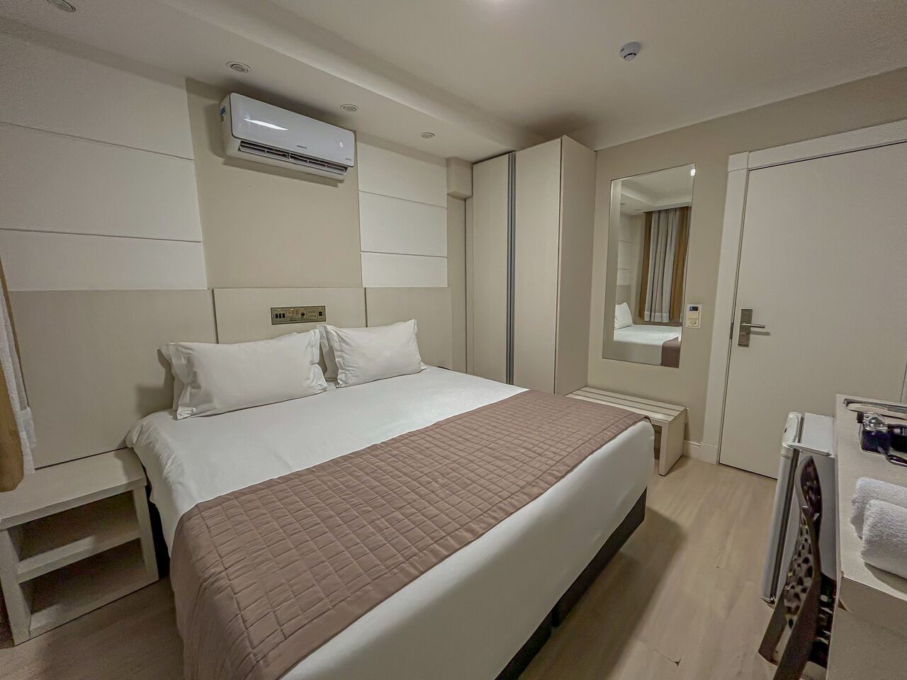 A hotel room with a bed, a bedside table, a mirror, and a bathroom with a towel rack.